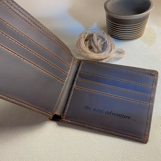 Brown leather wallet with 'the next adventure...' text on a light surface with yarn and cups in the background.