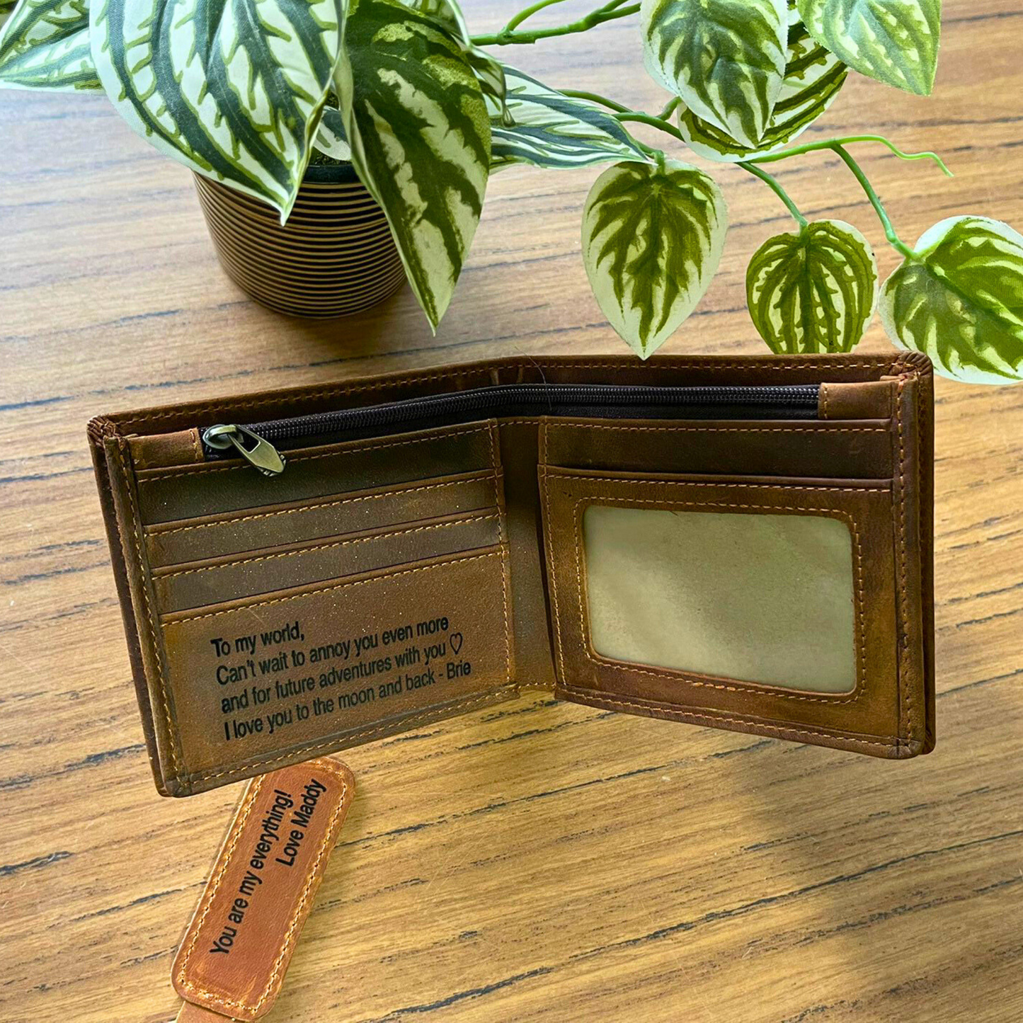 Brown leather wallet with engraved message on a wooden surface with plants in the background