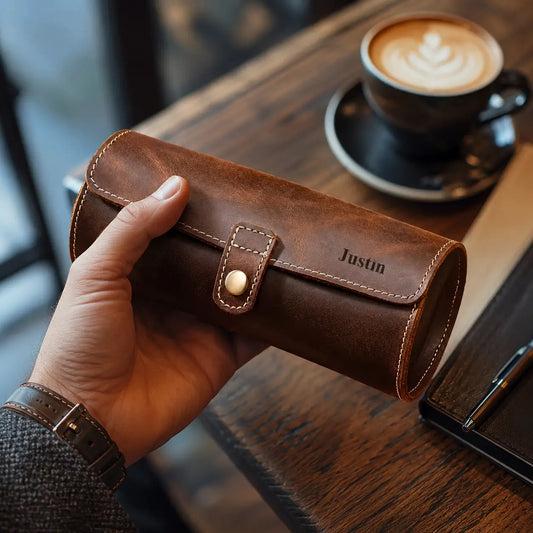 Close-up lifestyle image of a personalized brown leather watch roll case held in a man’s hand above a wooden café table beside a blurred latte cup. Warm natural lighting highlights the vintage leather texture, beige stitching and brass button closure.