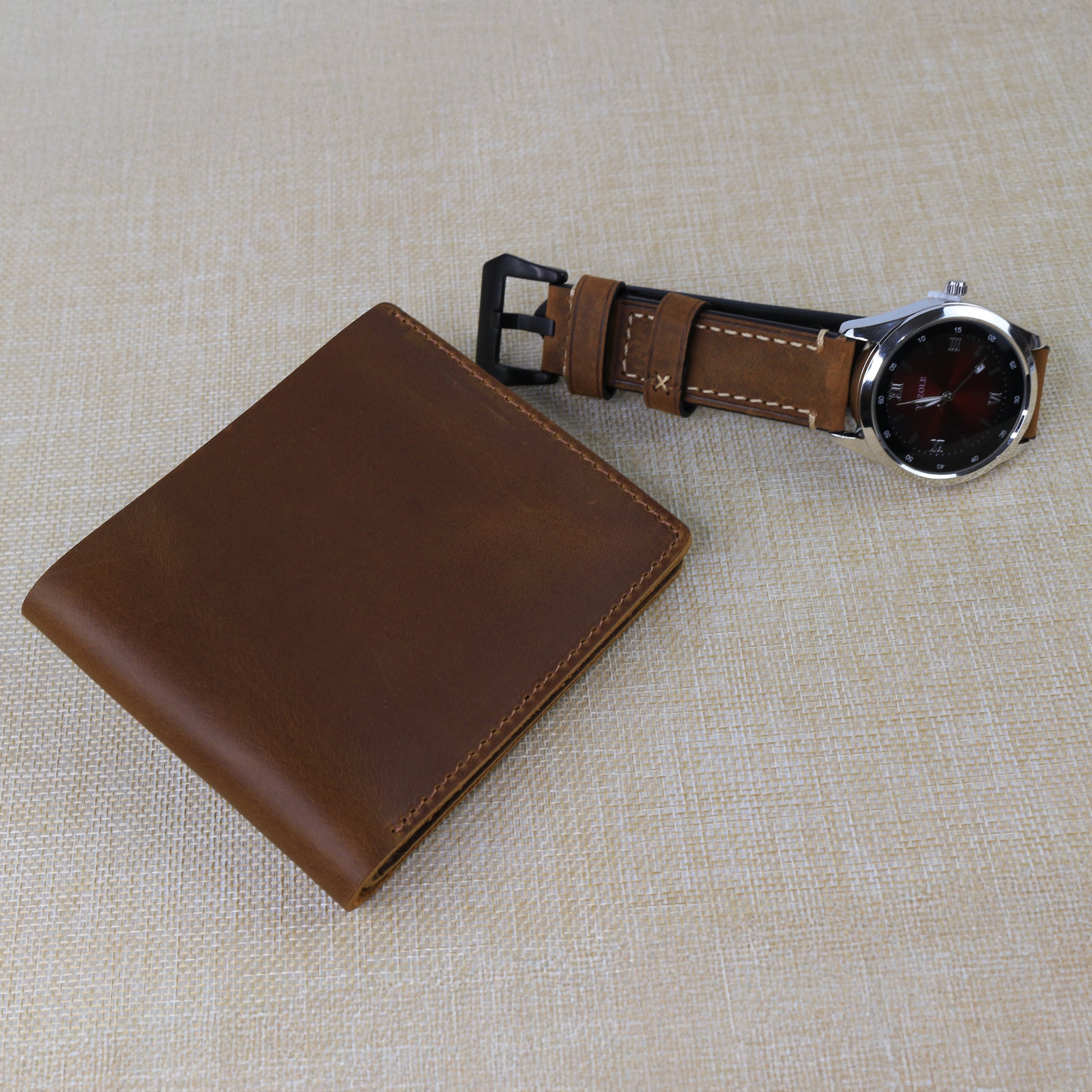 Personalized dark brown leather bifold wallet with the name "Royan" custom engraved on the front, shown with a matching leather watch strap on a textured surface.