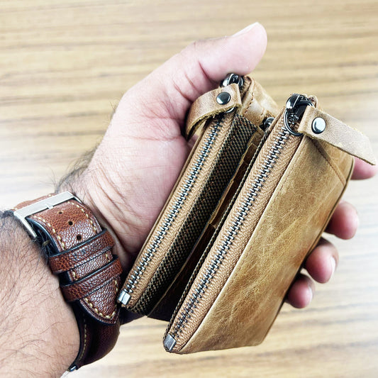 Hand holding a brown leather wallet with multiple zippers on a wooden surface