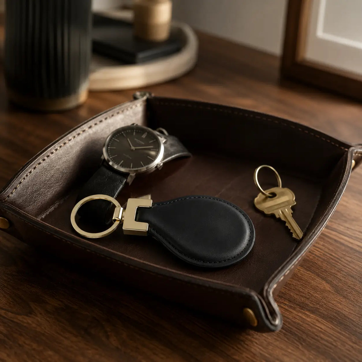 Black leather keychain with gold hardware styled beside wristwatch and keys on a black valet tray