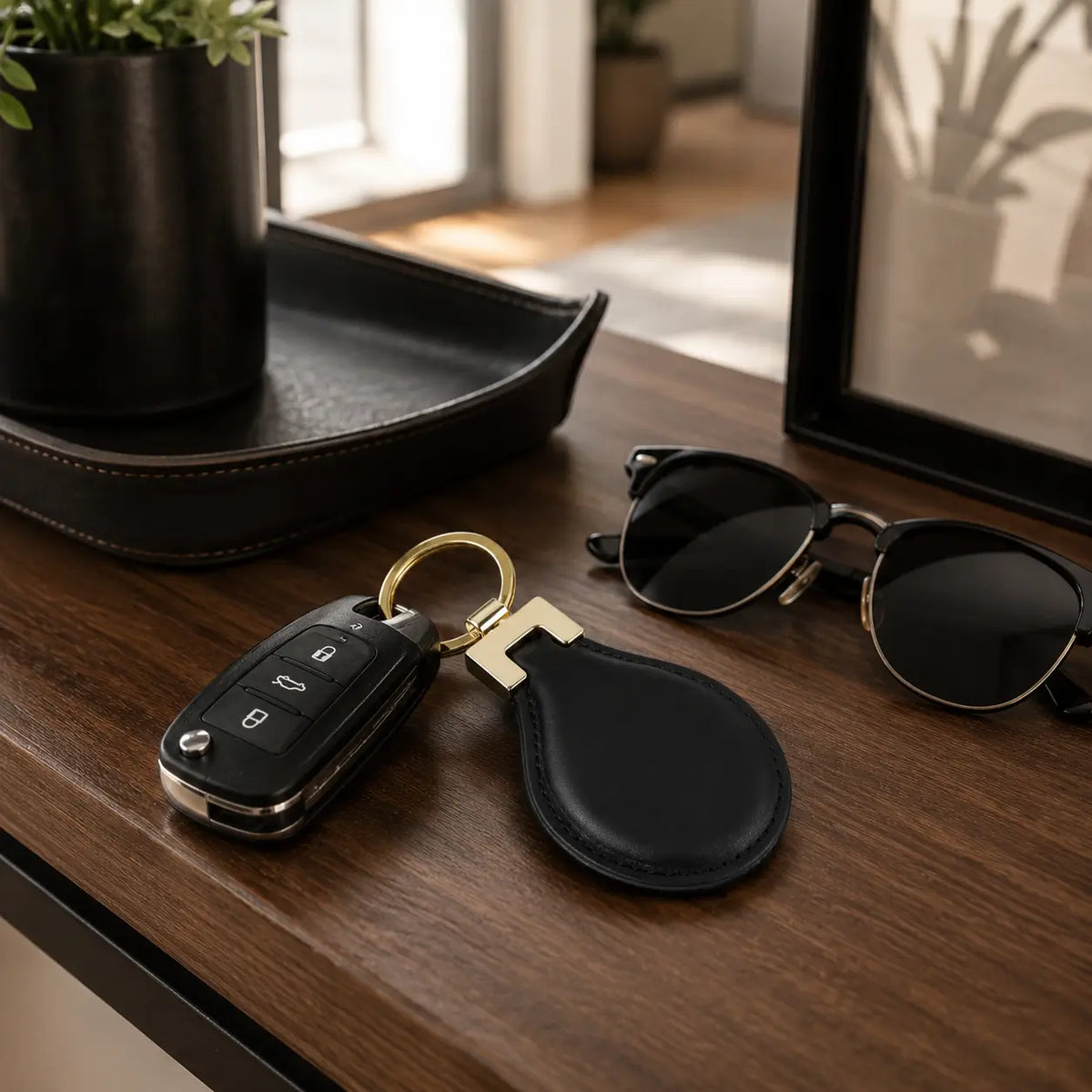 Black leather keychain displayed with car keys and sunglasses on a wooden desk in a modern home setting