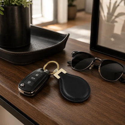 Black leather keychain displayed with car keys and sunglasses on a wooden desk in a modern home setting