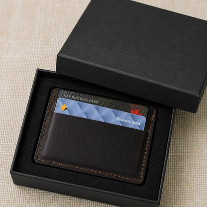Slim handmade leather credit card sleeve in a sleek black matte presentation box on a textured cream surface.