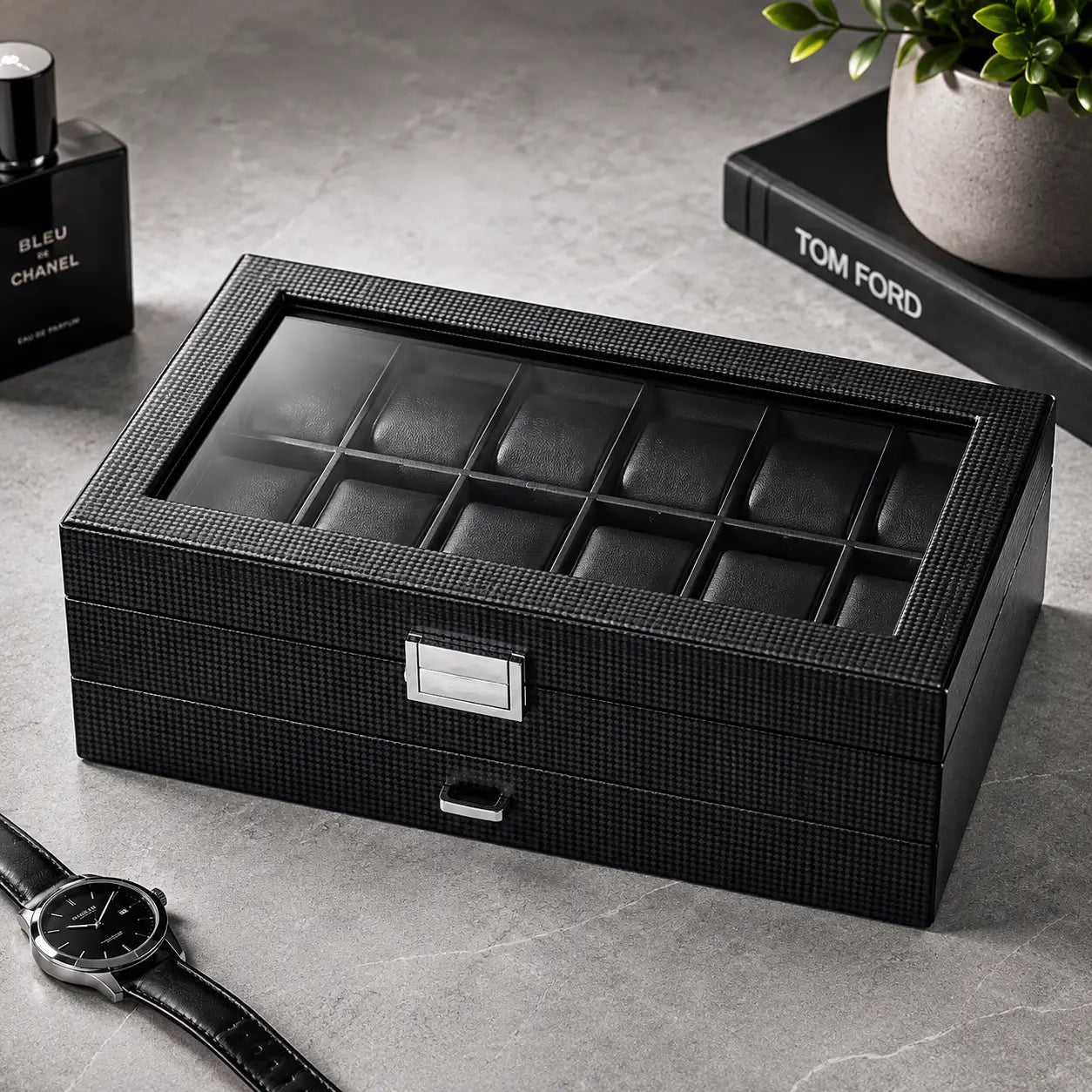 Closed carbon fiber men’s watch box with glass display lid and silver clasp placed on a modern grey stone surface beside luxury accessories, designer fragrance, and minimalist masculine décor.