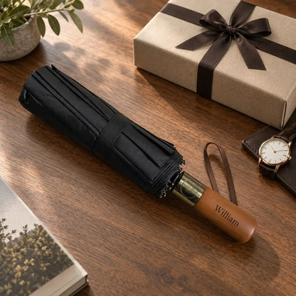 Personalised black folding umbrella with wooden handle engraved William beside gift box on wooden table