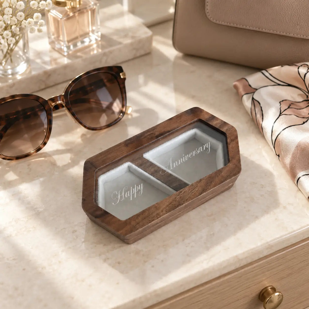 Personalised wooden ring box with engraved glass or lid displayed beside sunglasses and a handbag on a neutral dressing table.