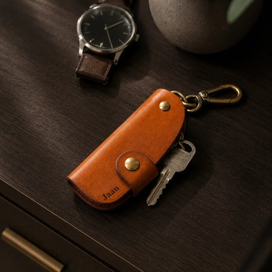 Personalised tan leather key holder engraved with James, featuring brass snap closure and metal key hook beside wristwatch