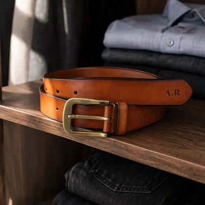 Alt text: Personalized brown leather belt with engraved initials and antique brass buckle displayed on a wooden shelf beside folded shirts and denim, elegant lifestyle product photography.