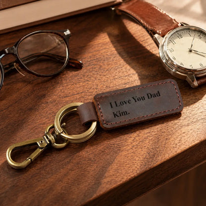 Personalised brown leather keychain engraved with I Love You Dad and Kim, featuring antique brass hardware beside watch and glasses