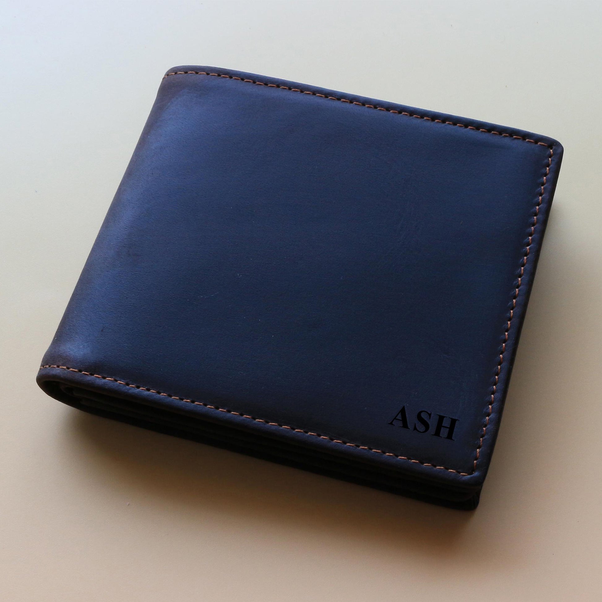 Dark brown leather wallet with 'ASH' monogram engraved on it on a beige background