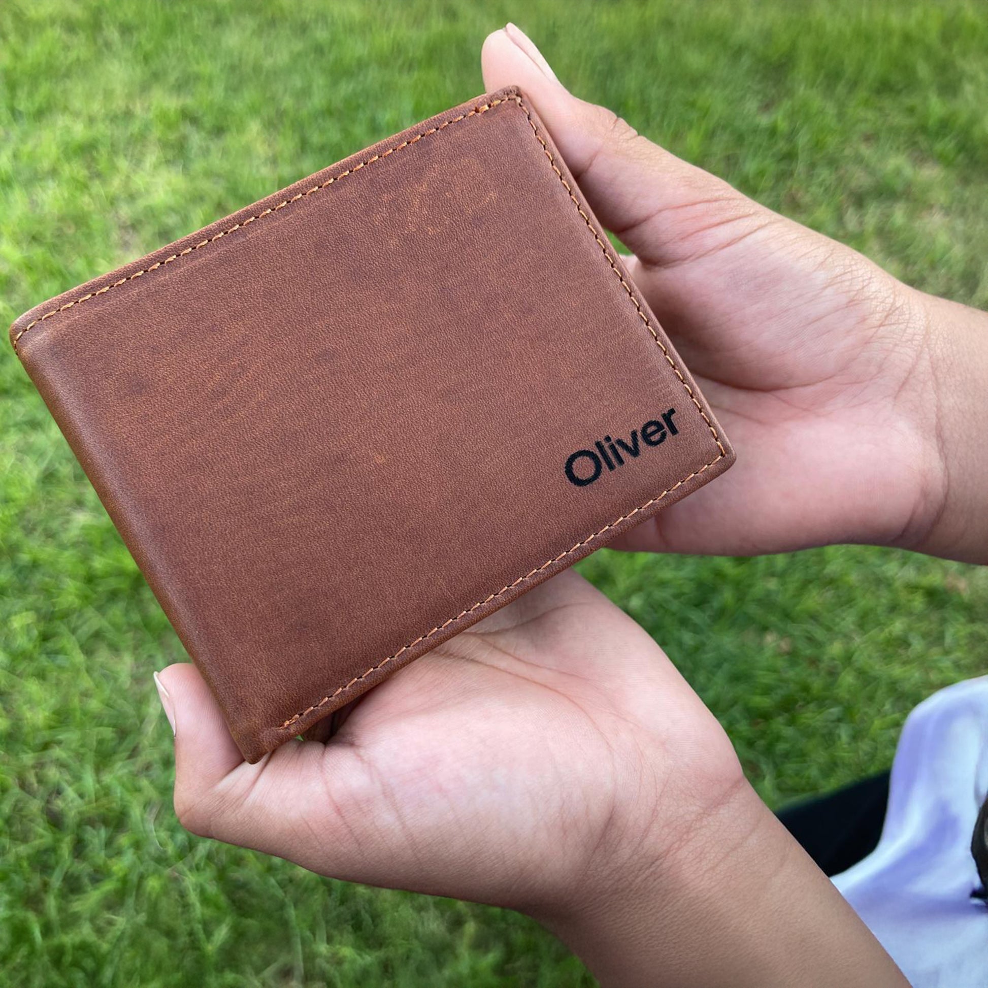 Brown leather wallet with 'Oliver' branding held in a hand against a grassy background
