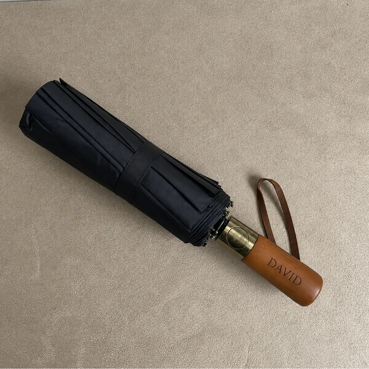 Black compact folding umbrella with wooden handle and brown wrist strap on neutral background
