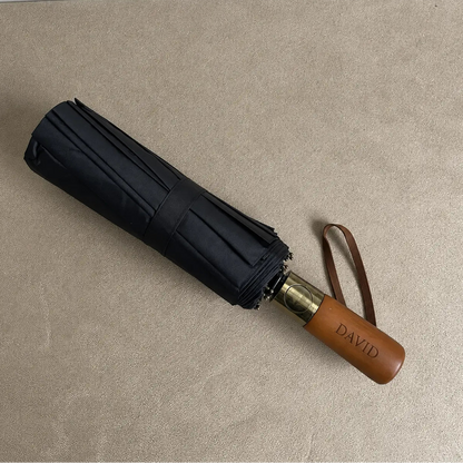 Black compact folding umbrella with wooden handle and brown wrist strap on neutral background