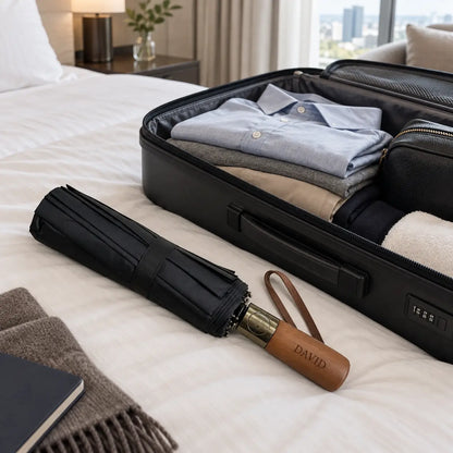 Personalised travel umbrella with black canopy and wooden handle engraved David beside packed suitcase on hotel bed