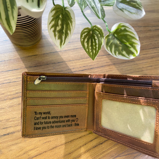 Open brown leather wallet with engraved message on a wooden surface with plants in the background