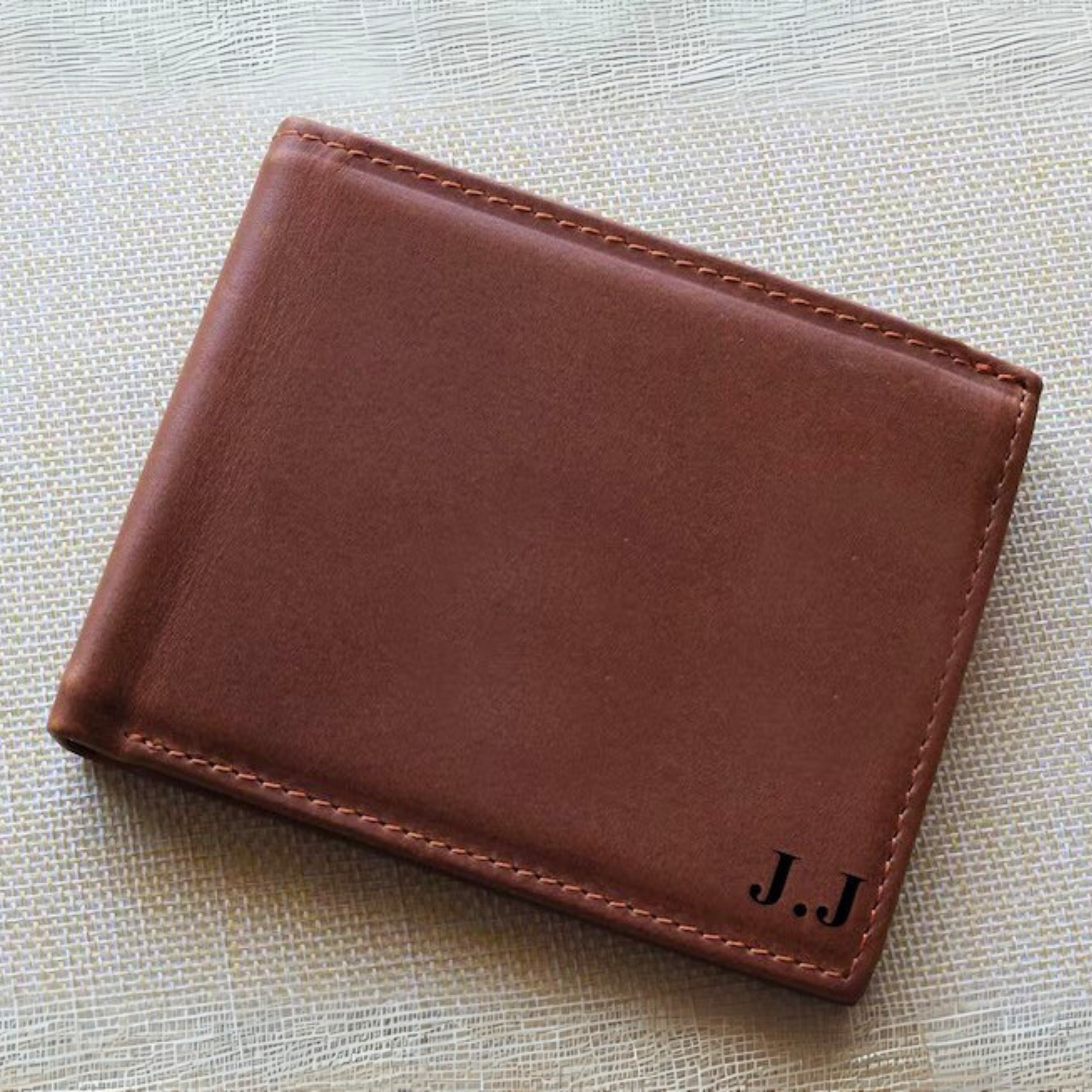 Brown leather wallet with initials 'J.J' engraved it, on a textured beige surface.