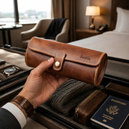 Luxury travel lifestyle image of a personalized tan-brown leather watch roll case held above an open suitcase inside a modern hotel room. Soft ambient lighting emphasizes the smooth leather finish, handcrafted stitching and premium travel aesthetic.