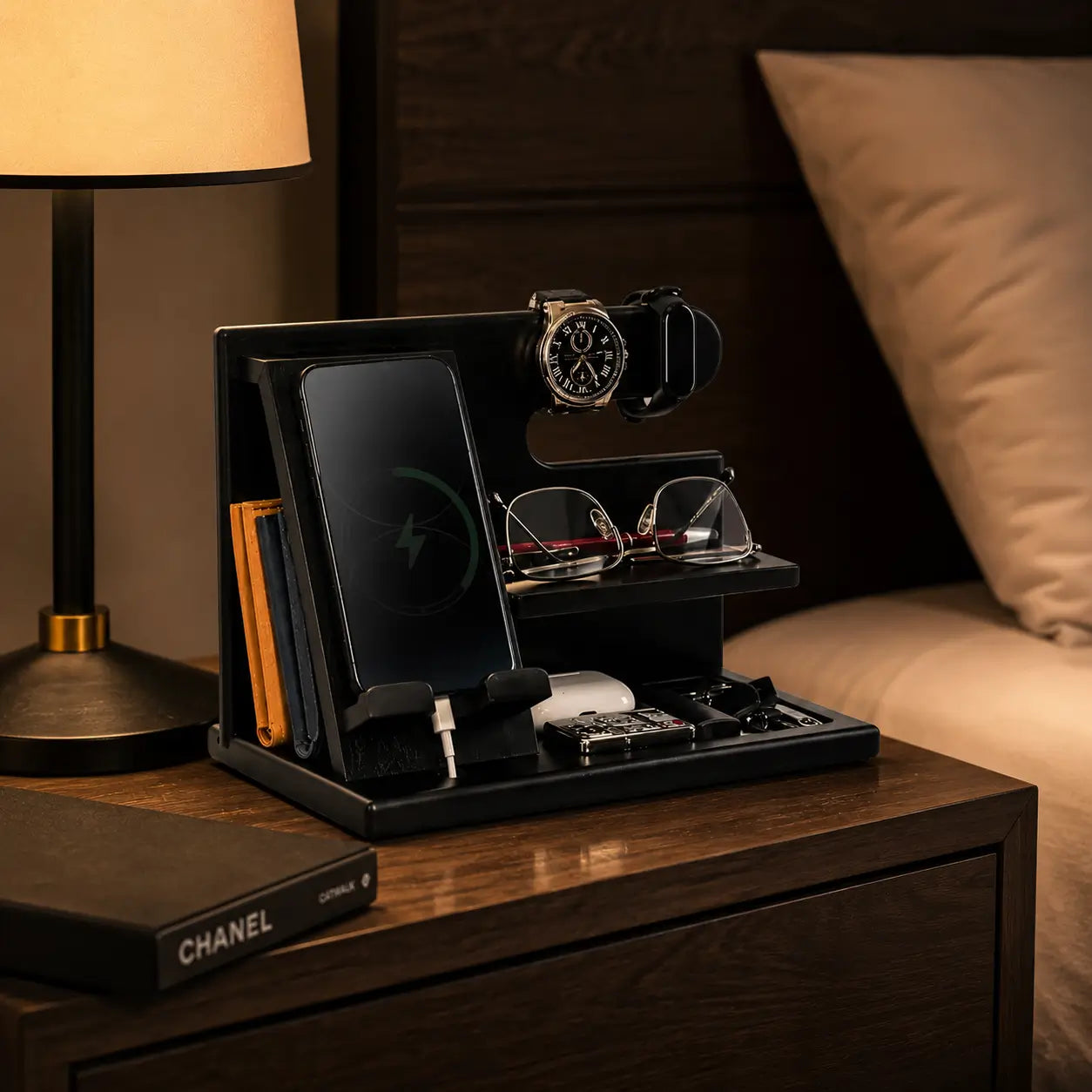 Black wooden charging station organizer placed on a modern bedside table with warm lamp lighting, featuring smartphone charging, watch holder and neatly organized everyday accessories.