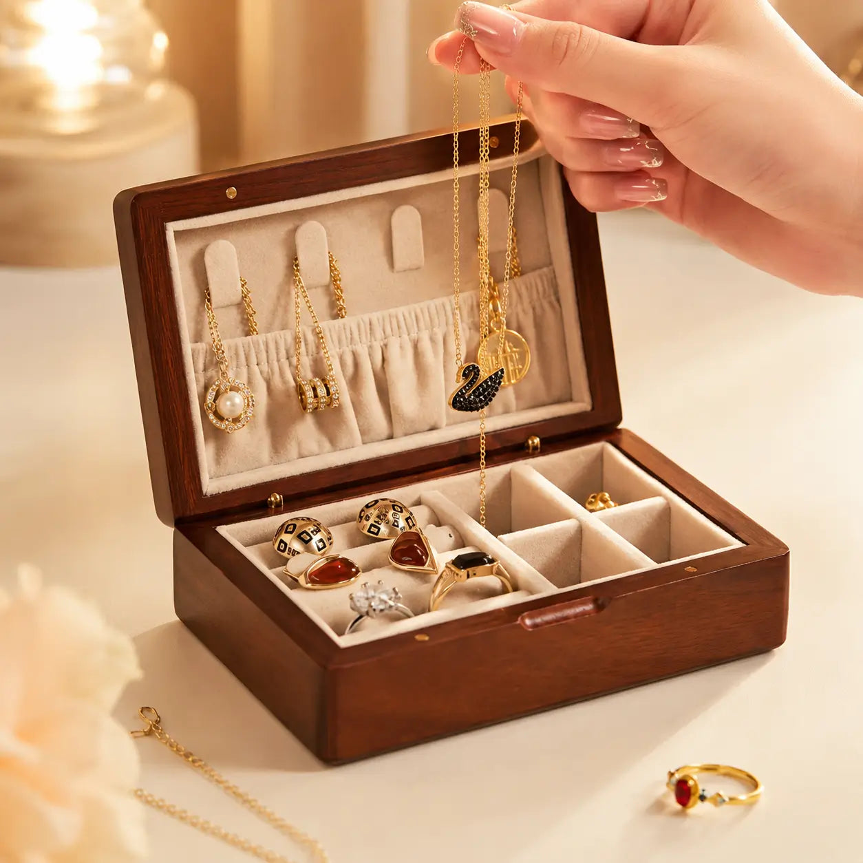 Close-up of a walnut wooden jewelry organizer with beige velvet lining while a feminine hand holds a delicate gold necklace above neatly arranged rings and pendants.